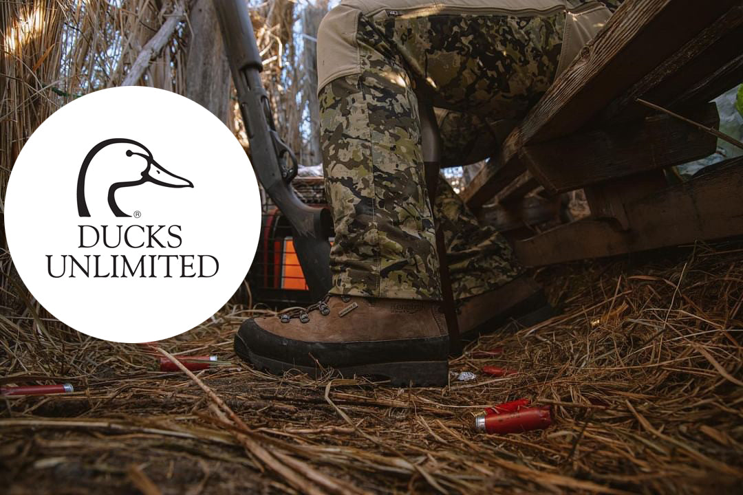 A person wearing camouflage hunting pants and brown boots sits under a wooden structure in a wooded setting, with a shotgun resting beside them. Red shotgun shells are scattered on the straw ground. White Ducks Unlimited Logo with black profile of duck head.